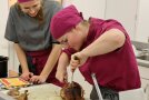 Zwei Frauen in einheitlicher Arbeitskleidung tranchieren eine gebackene Weihnachtsente.