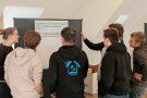 Mehrere Studierende stehen im Halbkreis mit Rücken zur Kamera vor einem Flipchart.