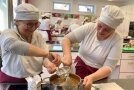 Zwei Frauen in einheitlichem Küchengewand beim Rühren einer süßen Masse in einem Topf. 