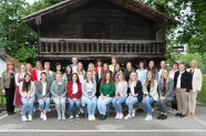 Personengruppe steht und sitzt für ein Gruppenbild in zwei Reihen vor einer Holzhütte.