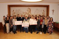 Große Gruppe von Menschen in Saal, drei davon präsentieren große Spendenschecks in die Kamera.