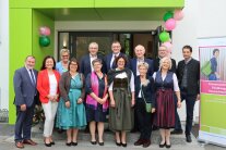 Gruppe von festlich gekleideten Personen in zwei Reihen vor Gebäudeeingang, rechts steht eine Informationstafel.