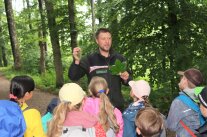 Mann vor Gruppe mit Kindern hebt Blatt hoch und erklärt.