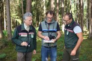 Drei Männer im Wald schauen auf ein Dokument, das der Mann in der Mitte hält.