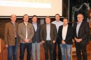 Sieben Männer in Jackets und Hemden stehen in Reihe für ein Gruppenbild, dahinter Projektionsfläche.