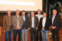 Sieben Männer in Jackets und Hemden stehen in Reihe für ein Gruppenbild, dahinter Projektionsfläche.