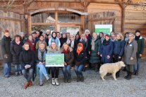 Personengruppe sitzend und stehend mit Tafel und Hund vor hölzernem Gebäude.