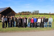 Große Personengruppe steht in REihe am Rand einer Wiese in der Sonne, im Hintergrund Scheune, Feld und Waldsaum.